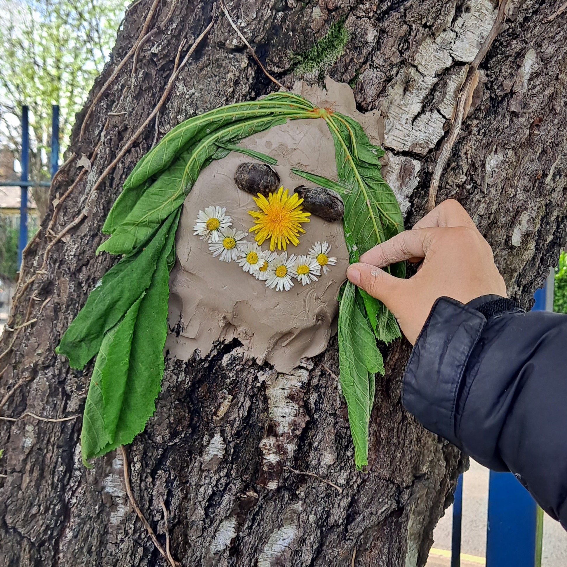 Wyneb clai ar goeden gyda dail gwyrdd ar gyfer gwallt, dant y llew ar gyfer y trwyn a llygaid yr haul ar gyfer gwên.